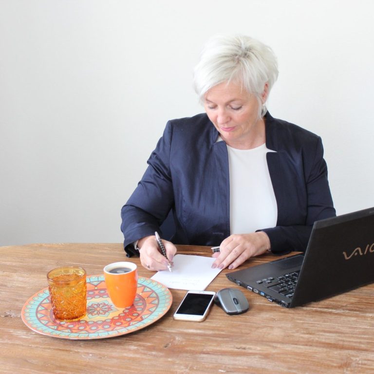 Eine Frau schreibt am Tisch, neben ihr ein Laptop, Tasse und Smartphone.