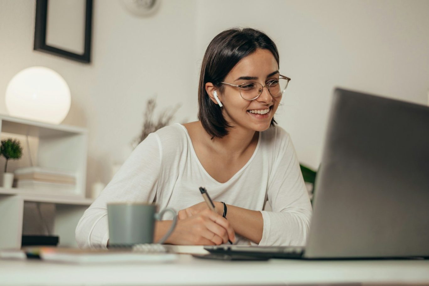 Lächelnde Frau mit Brille, die an einem Laptop arbeitet und einen Kaffeebecher hält.