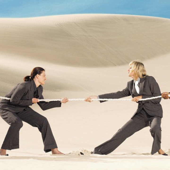 Zwei Frauen in Anzügen ziehen an einem Seil in einer Wüstenlandschaft.