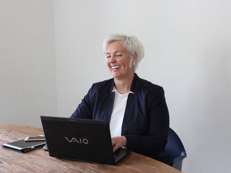 Webinarreihe 2 Lächelnde Frau mit weißem Haar sitzt am Tisch und arbeitet an einem Laptop.