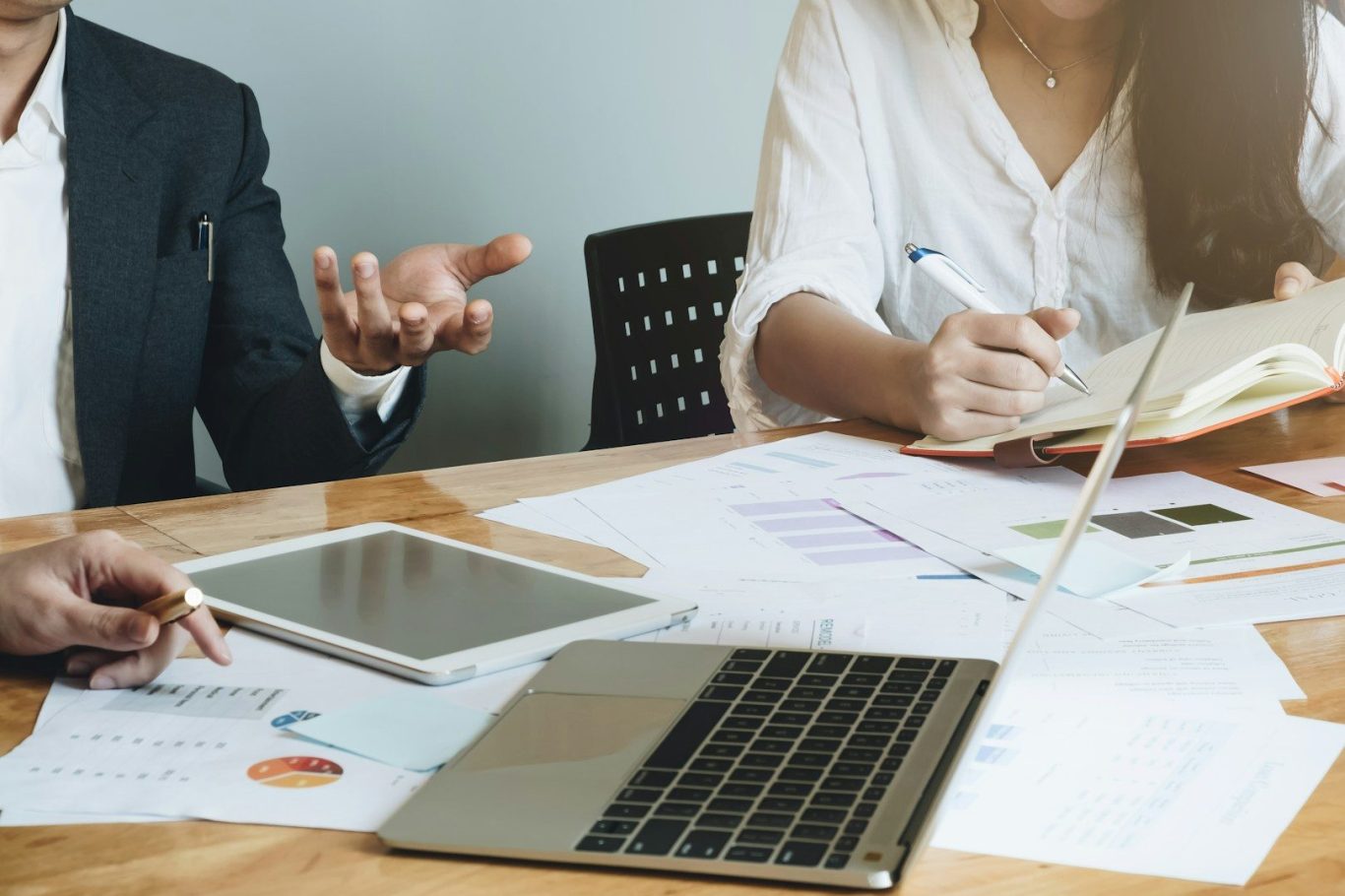 Zwei Personen diskutieren am Tisch, Laptops und Unterlagen sind sichtbar.