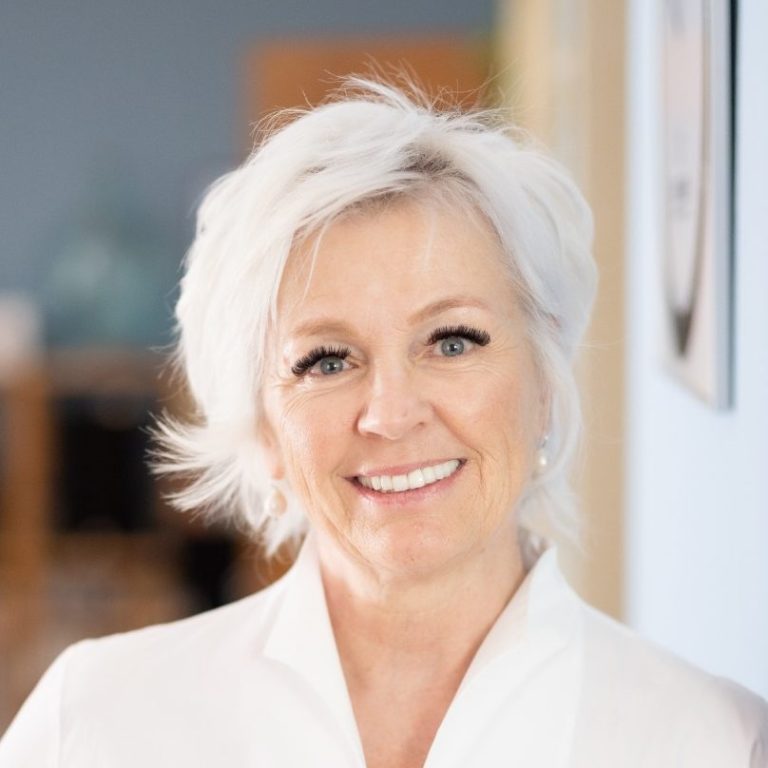 Lächelnde Frau mit kurzen, grauen Haaren und weißer Bluse, auf neutralem Hintergrund.