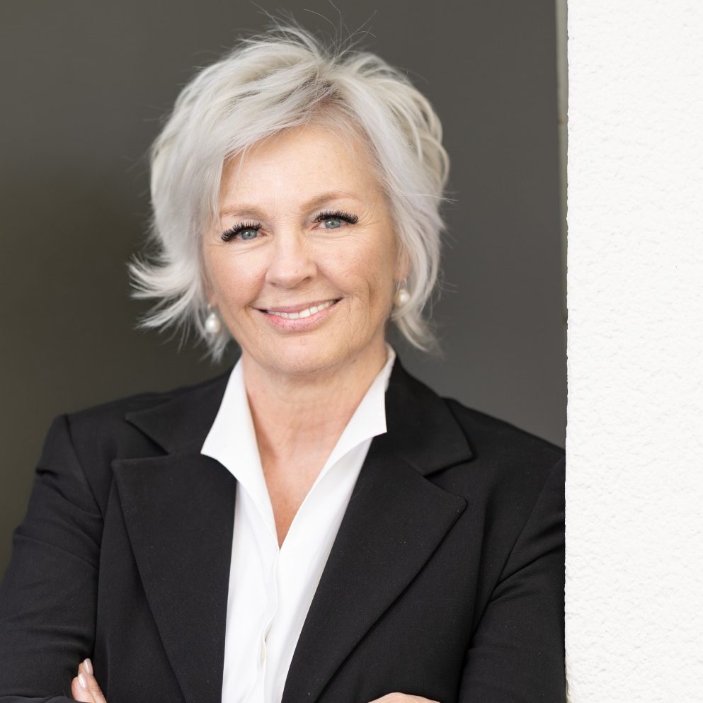 Porträt einer lächelnden älteren Frau mit grauen Haaren in einem hellen Hemd.